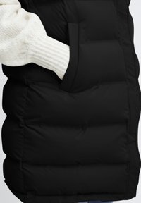 Crna puffer prsluk s bočnom džepom, koji ima kviltanu teksturu i rebrastu, off-white rukav koji proviri iz džepa.