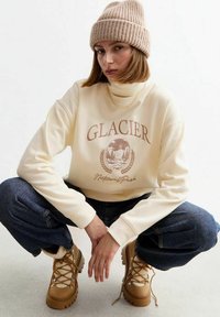 Sudadera crema con estampado de "Parque Nacional Glacier", combinada con jeans de pierna ancha de color azul oscuro y botas de cordones color topo con forro de piel sintética.