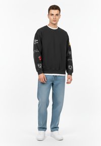 Felpa nera con grafiche sulle maniche, collo rotondo e polsini a coste, abbinata a jeans azzurri chiari e sneakers bianche.