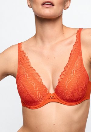 Woman wearing an orange lace bra with scalloped edges and underwire, showing from shoulders to midriff against a plain background.