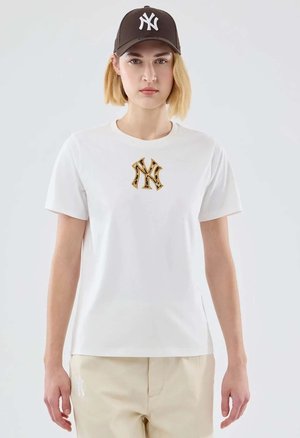 Junge Frau trägt weißes T-Shirt mit goldenem und schwarzem NY-Logo, beige Hose mit weißem NY-Logo und dunkelbraune NY-Baseballkappe, steht nach vorne.