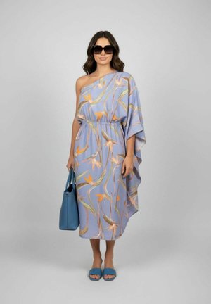 Femme portant une robe lavande à fleurs à épaules dénudées, de grandes lunettes de soleil noires, tenant un sac à main bleu, avec des sandales ouvertes assorties bleues, debout devant un fond gris.