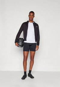 Must jalgratturi jakk valge t-särgi, mustade lühikeste pükste ja mustade jalgrattakingade peal. Must kiiver käes. Minimalistlik disain, vormitud lõige.