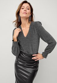 Sort, tætsiddende nederdel i faux læder parret med en mørkegrå bluse med glimmer, der har lange ærmer og en dyb V-hals.