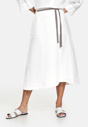 Vrouw draagt een witte midirok met een zwart-witte geweven riem en witte platte sandalen, staat met gekruiste benen.