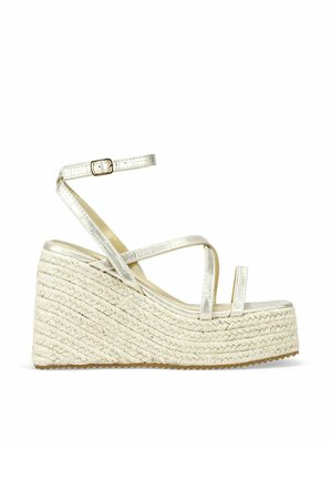 Sandalia cuña de color plata metálico con tiras entrelazadas y hebilla ajustable en el tobillo. Plataforma de cuerda texturizada y suela de goma.