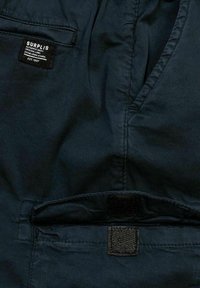 Pantaloni in tessuto scuro con una superficie liscia, dotati di un'etichetta in vita e un dettaglio di chiusura in Velcro sulla tasca anteriore.