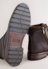 Bottes cheville en cuir marron avec accents texturés, semelle en caoutchouc avec rainures antidérapantes, et une fermeture éclair latérale. Doublure douce visible en haut.