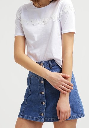 Vrouw die een wit T-shirt draagt met kanten hartmotieven over de borst en mouwen, gecombineerd met een blauwe spijkerrok met knopen aan de voorkant.