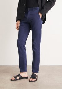 Chino din bumbac de culoare bleumarin, cu croi slim, dotat cu buzunare laterale și închidere cu buton, asociat cu sandale negre texturate cu talpă de tip slide.