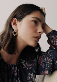Gold heart-shaped earrings with a textured, ribbed design. The wearer has long brown hair and is dressed in a floral-patterned blouse.