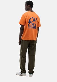 Homme portant un t-shirt orange avec un graphisme de camping bleu et le texte "VÊTEMENTS DE PLEIN AIR", un pantalon vert olive et des baskets blanches, se tenant de profil.