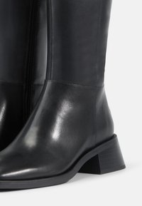 Μαύρες δερμάτινες μπότες ως το γόνατο με λεία επιφάνεια, πλευρικό φερμουάρ και ταπεινό τακούνι. Διαθέτουν στρογγυλή μύτη και κομψή σιλουέτα.