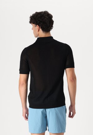 Polo shirt tricotat negru cu un model texturat și respirabil. Mâneci scurte și guler clasic. Purtați cu pantaloni scurți de culoare albastru deschis.