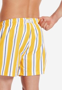 Costume da bagno giallo con strisce verticali bianche e blu, cintura elastica e un'etichetta sul retro. Tessuto liscio e a asciugatura rapida.