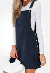 Vestido de tirantes de pana azul marino con un bolsillo frontal, botones laterales y un corte relajado, combinado con un bolso blanco.