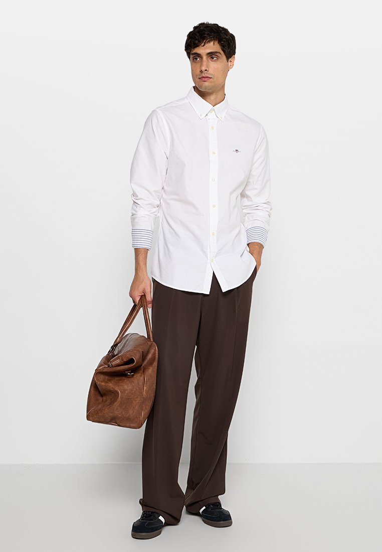 Weißes Hemd mit Knöpfen und gestreiften Bündchen, braune lässige Hose im Relaxed-Fit-Stil und eine braune Leder-Reisetasche. Legere Schuhe runden den Look ab.