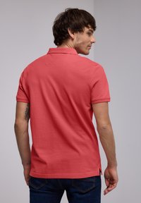 Polo corail en coton, avec un col classique et des manches courtes. L'arrière comprend une étiquette et des fentes sur les côtés à l'ourlet.