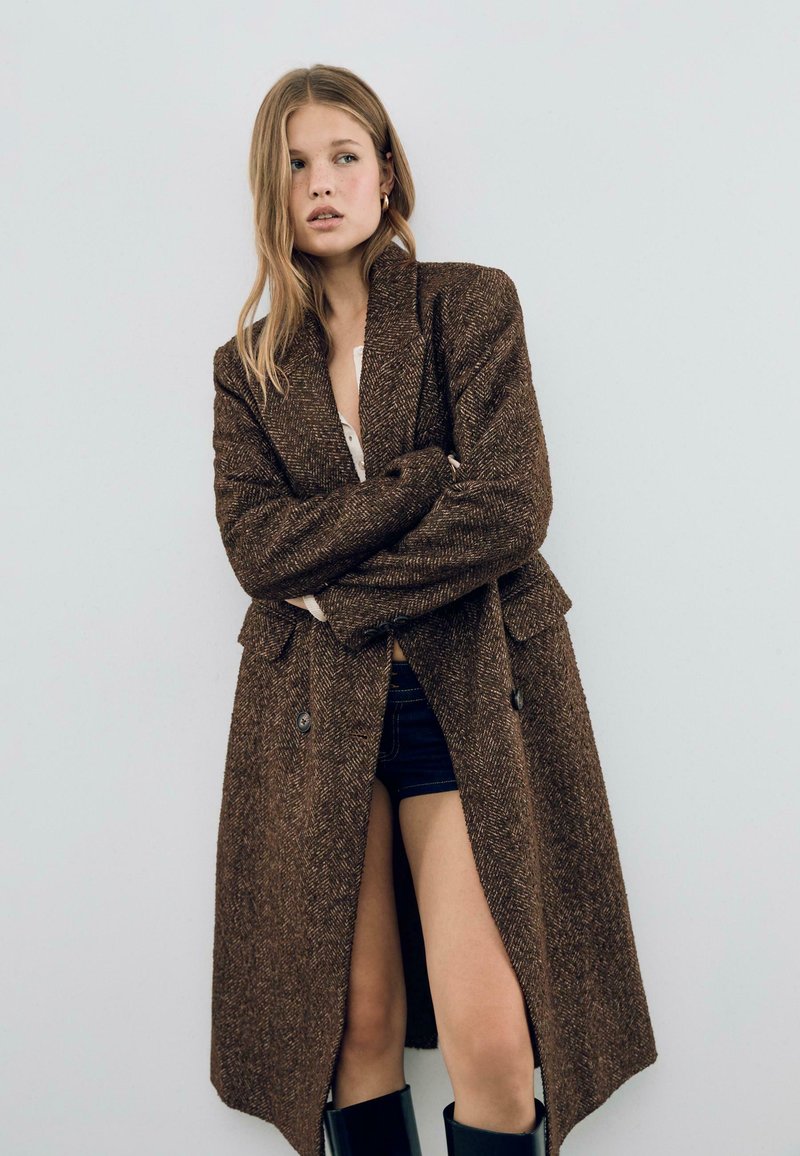 Manteau texturé marron avec un design long, revers crantés et grands boutons ; porté sur une chemise blanche et un short en denim, assorti à des bottes noires.