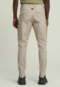 Pantaloni cargo beige con un design aderente, dotati di quattro tasche posteriori, cerniere laterali e tessuto testurizzato. Abbinati a calzature nere.