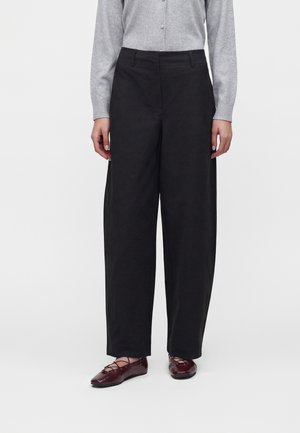 Personne debout portant un pantalon noir large taille haute, un haut gris à manches longues boutonné, et des ballerines bordeaux avec des brides croisées.