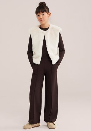 Jeune femme debout, les mains dans les poches, portant une veste en fourrure blanche sur un haut à manches longues foncé et un pantalon large assorti avec des baskets.