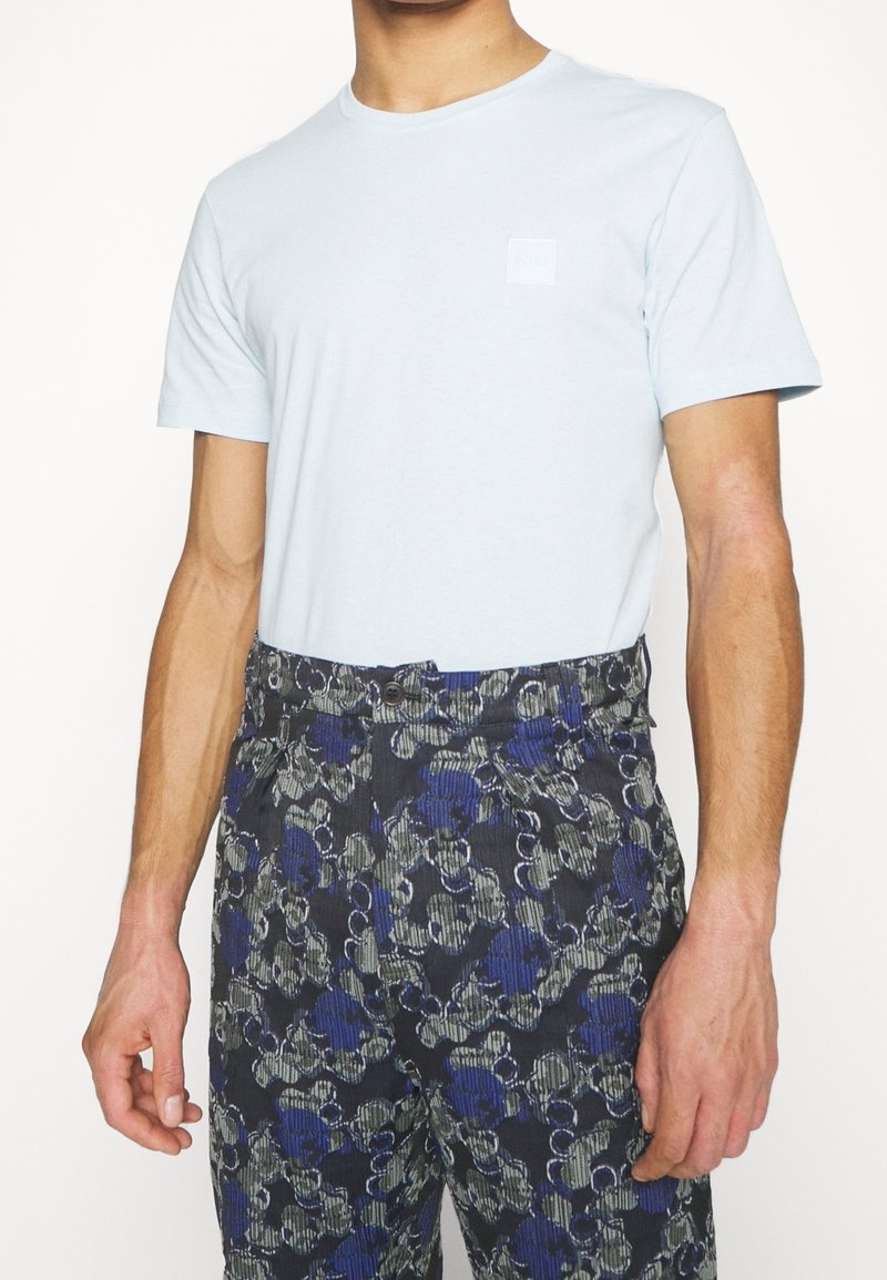 T-shirt en coton bleu clair avec un col ras du cou. Associé à un pantalon sombre à motifs avec des dessins floraux bleus, verts et gris et une fermeture à boutons.