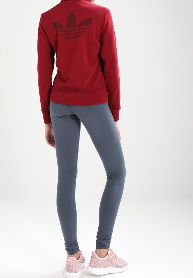 En person som bär en röd huvtröja med en stor logotyp på ryggen, grå leggings och rosa träningsskor, stående mot en enkel bakgrund.