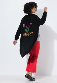 Femme souriante, portant un cardigan à capuche noir avec "MORE AMORE" et un smiley dans le dos, un pantalon rouge et des chaussures noires à clous, posant contre un mur blanc.