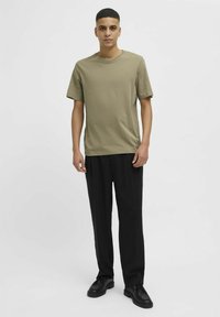 Camiseta de manga corta color verde oliva con escote redondo, combinada con pantalones negros de pierna ancha y zapatos negros con cordones, sobre un fondo blanco.