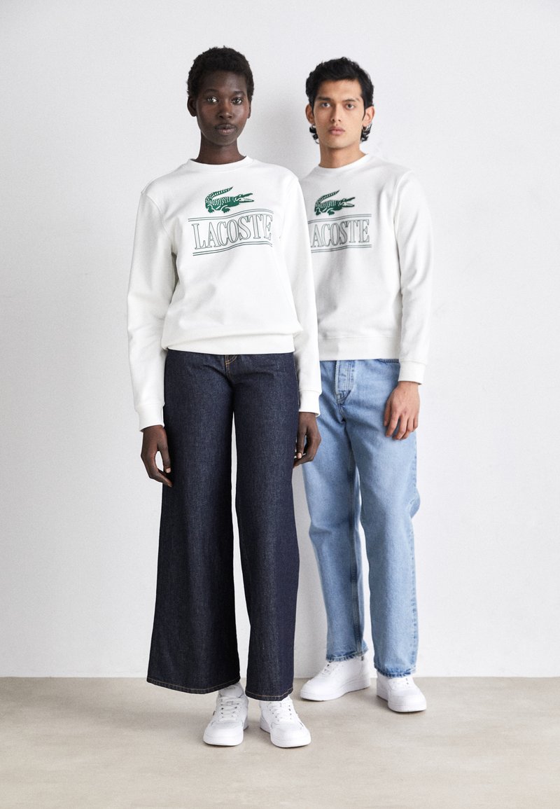 Sudaderas blancas con logo verde de Lacoste, pantalones de mezclilla oscuros de pierna ancha, jeans de color azul claro y zapatillas blancas. Texturas suaves, corte casual.