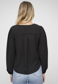 Blusa negra con mangas largas abullonadas y cuello redondo. Tejido transparente con costura recta en la espalda y detalle sutil de pliegue trasero.
