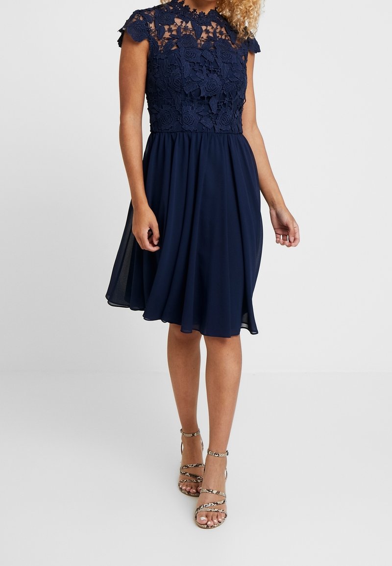 Robe bleu marine avec un corsage en dentelle florale et des manches courtes à volants. Jupe fluide en tissu léger, tombant juste au-dessus des genoux.