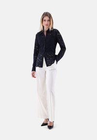 Camicia a maniche lunghe in pizzo blu navy con apertura frontale a bottoni, abbinata a pantaloni bianchi a gamba larga e décolleté neri con punta a punta.