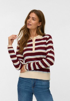 Vrouw met lang haar die een maroon, beige en lichtblauw gestreepte lange mouwen polo draagt en blauwe jeans, terwijl ze haar haar aanraakt.
