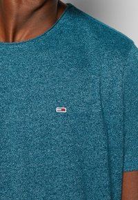 Tyrkysové tričko s krátkým rukávem vyrobené z texturované tkaniny, s malou nášivkou loga v červené, bílé a modré barvě na levé části hrudi.