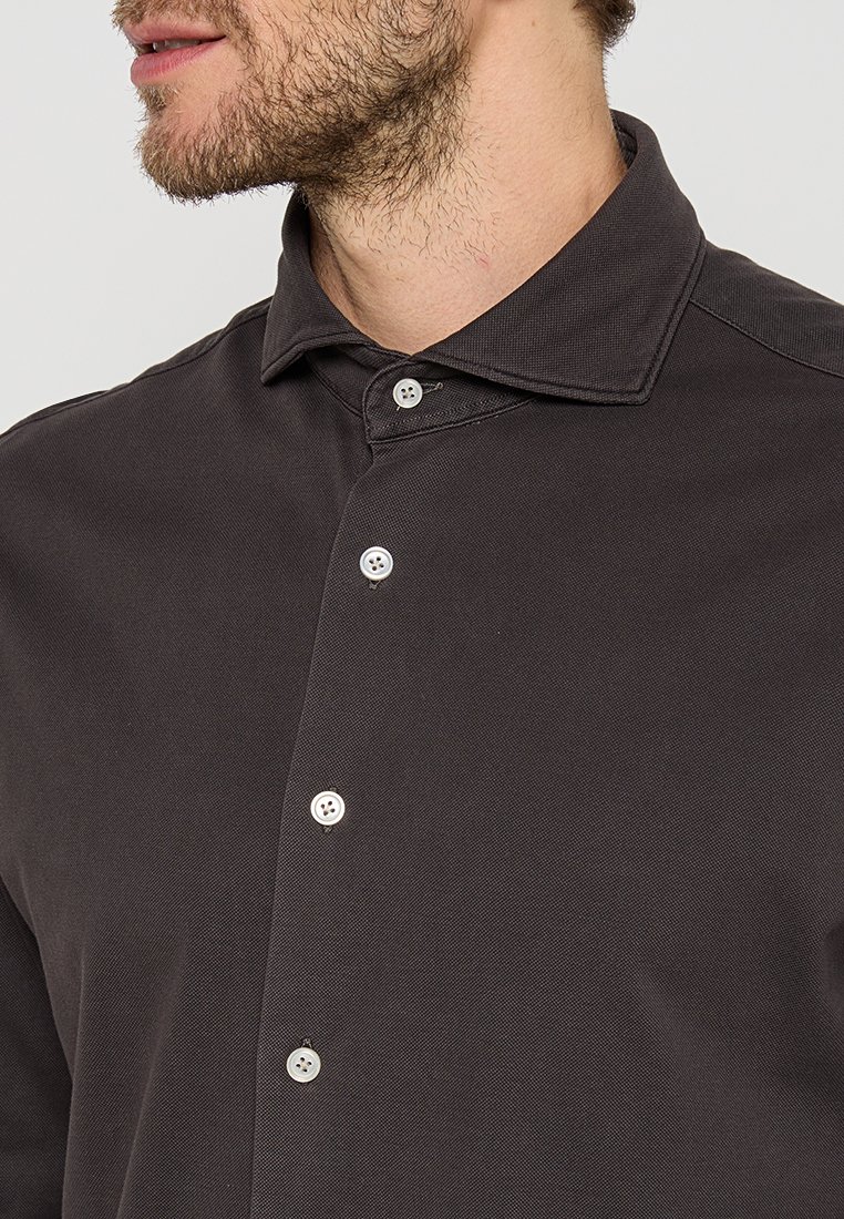 Man met een lichte baard, gekleed in een donkerbruine gekraagde shirt met vier zichtbare witte knopen, op een effen achtergrond.