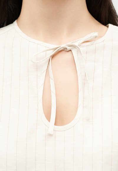 Κοντινό πλάνο ενός κρεμ μπλουζάκι με λεπτές κάθετες ρίγες, που διαθέτει λαιμόκοψη με άνοιγμα σε σχήμα κλειδιού και έχει δεμένο φιόγκο από ύφασμα.