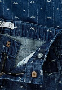 Denim teksad, kus on sinine kangas valgete lipsukestega, lukuga suletud ja nööbiga. Sisemine vöökoht näitab hooldusetiketti ja puuvillast tekstuuri.