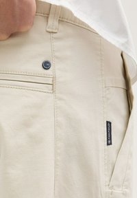 Beige katoenen broek met een gestructureerde afwerking, voorzien van een metalen knoop en een klein merklabel op de zijzak. Twee achterzakken zichtbaar.