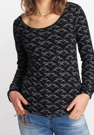 Frau trägt schwarzes Langarmshirt mit weißen Schleifenmustern, blaue Jeans und eine goldene Halskette, Hände teilweise in den Taschen.