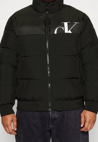 Sort dunjakke med høj krave, lynlås, ribstrikkede manchetter og "CK"-logo i hvid på brystet, med quiltet tekstur.