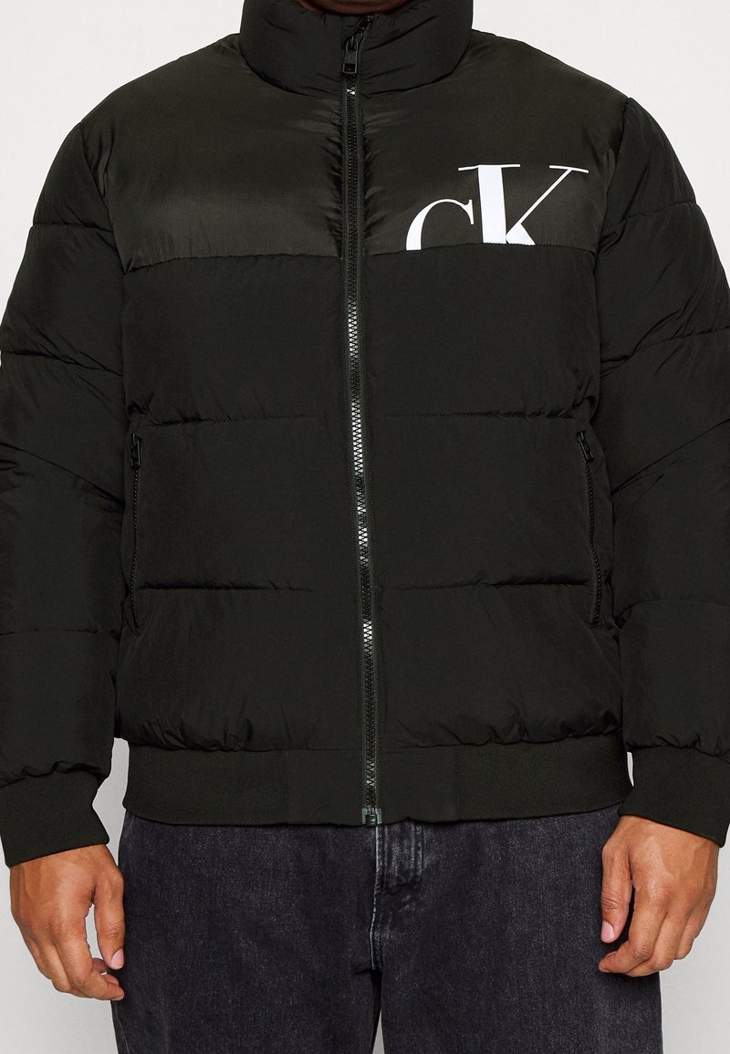 Sort dunjakke med høj krave, lynlås, ribstrikkede manchetter og "CK"-logo i hvid på brystet, med quiltet tekstur.