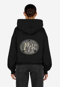 Juodas džemperis su dideliu apvaliu logotipu ant nugaros, turinčiu šviesios spalvos tekstą. Medžiaga atrodo minkšta, su laisvu apdailu.