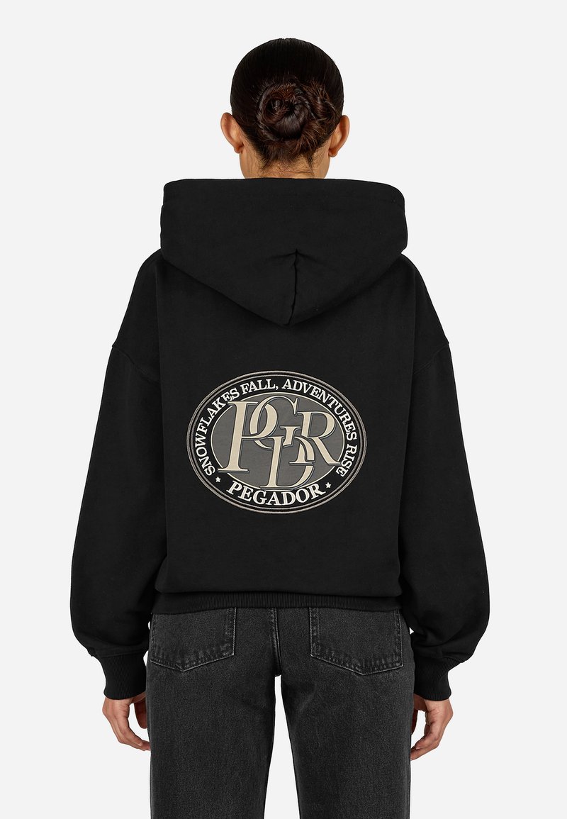 Juodas džemperis su dideliu apvaliu logotipu ant nugaros, turinčiu šviesios spalvos tekstą. Medžiaga atrodo minkšta, su laisvu apdailu.