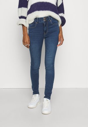 Osoba ubrana w ciemnoniebieskie obcisłe dżinsy, białe trampki oraz biało-fioletowy sweter w paski, wykonany na drutach.