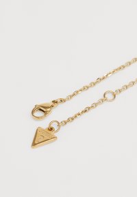 Collana in metallo color oro con pendente triangolare caratterizzato da un logo testurizzato. Include un gancio a bavaglino e dettagli di anelli circolari.