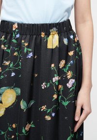 Μαύρη φούστα με ελαστική ζώνη μέσης που έχει λουλουδάτο και κίτρινο μοτίβο λεμονιού, φορεμένη με ανοιχτό μπλε τοπ και ένα ορατό χέρι να ακουμπάει στο πλάι.
