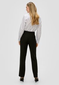 Zwarte op maat gemaakte broek met een rechte pijp, gedragen met een wit gestreept overhemd met knoopsluiting, voorzien van lange mouwen en een achterzakdetail.