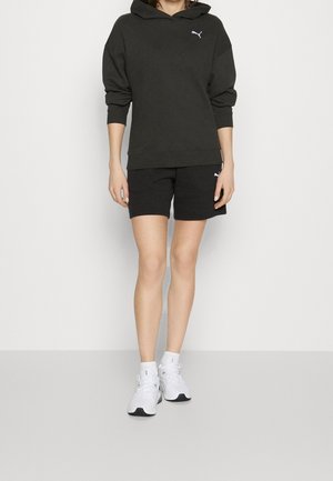 Personne portant un sweat à capuche Puma noir et un short avec des baskets blanches de sport devant un fond uni.